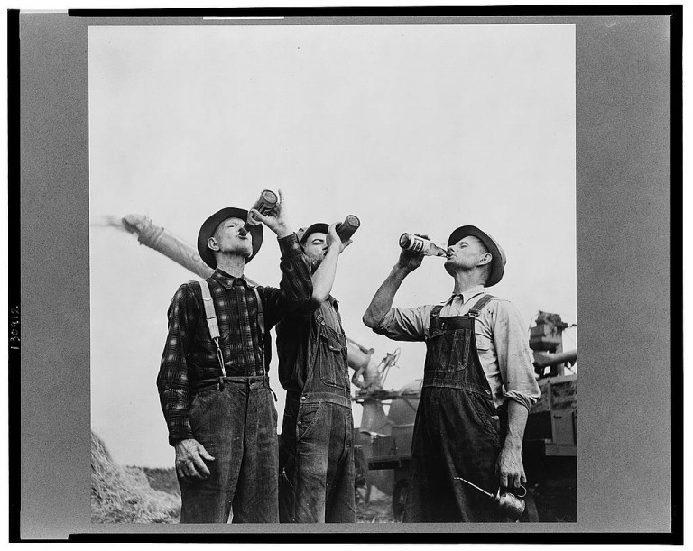 Cervejas feitas para o trabalhador: foto preto e branca antiga com três fazendeiros bebem suas cervejas no campo a frente de uma máquina colheitadeira antiga